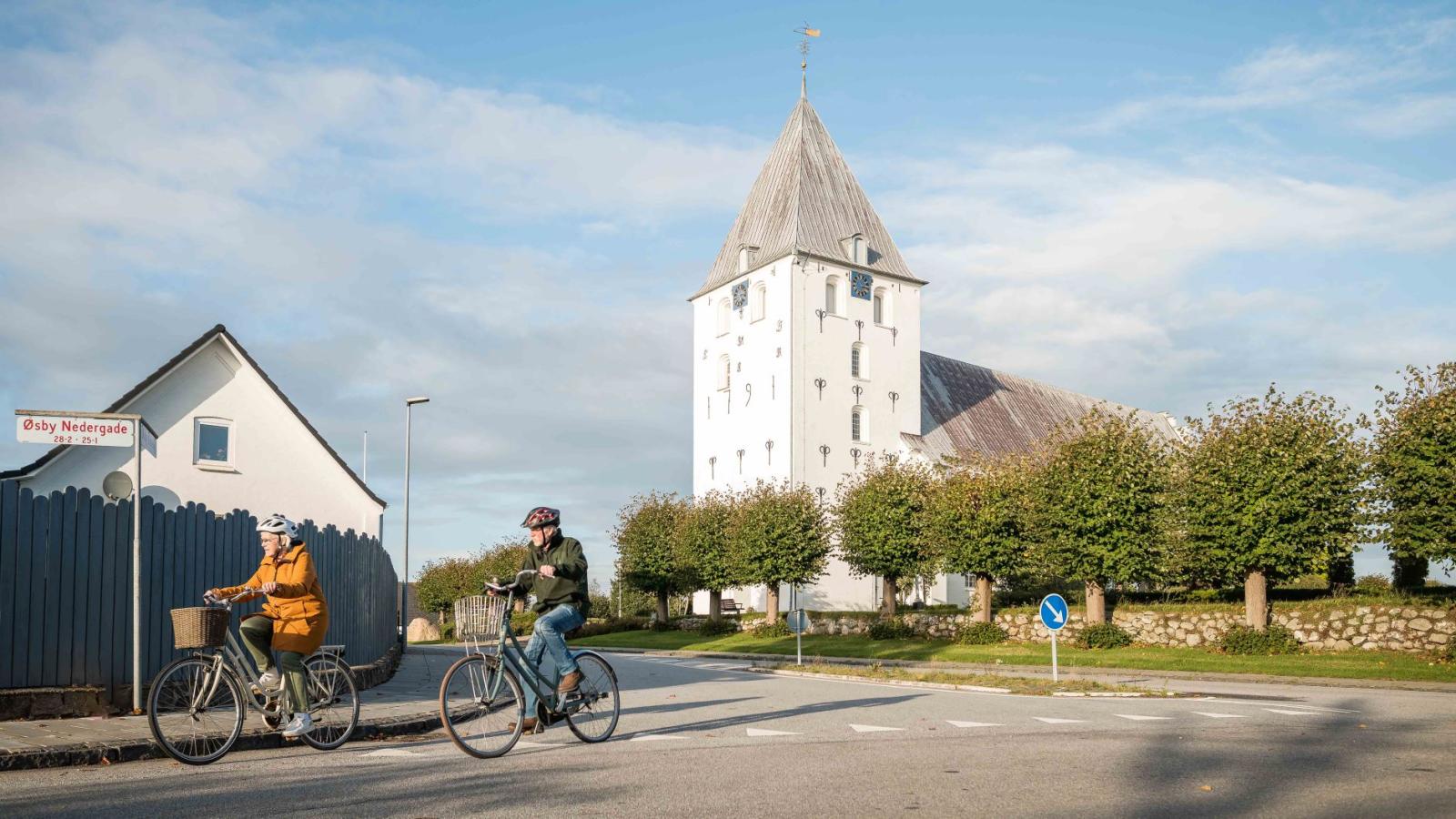 Et ældre par på cykeltur. I baggrunden kan man se en hvid kirke. 